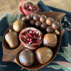 Puede incluir: Un cuenco de madera con forma de hoja lleno de frutas decorativas de madera y una flor roja y rosa. Las frutas de madera incluyen peras, uvas y otras formas redondas. El cuenco se encuentra sobre una tela con estampado floral.