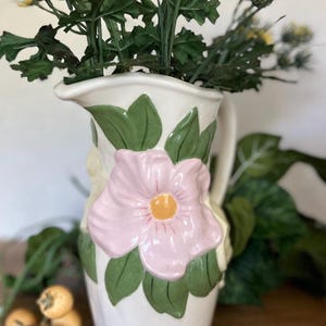 May include: A white ceramic pitcher with a pink flower and green leaves design. The pitcher is filled with green foliage and yellow flowers. The pitcher has a handle and a spout.