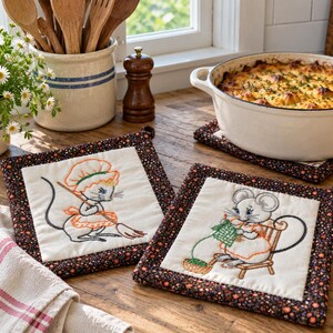 May include: Two square fabric pot holders with dark floral borders and cream-colored centers. One features an embroidered mouse wearing a chef hat and apron holding a broom. The other shows an embroidered mouse sitting on a chair knitting.