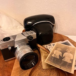 Puede incluir: Una cámara Kodak Instamatic Reflex vintage con cuerpo negro y plateado, lente y estuche negro. Junto a la cámara hay varias fotografías antiguas en blanco y negro sobre una bandeja de madera.