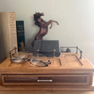 Peut inclure: Un organiseur de bureau en bois avec un tiroir, contenant des lunettes, un portefeuille gris foncé et une statue de cheval en bronze. L'organisateur a des accents en laiton et une finition en bois naturel, pour une esthétique classique.
