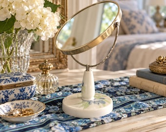 May include: A round tabletop mirror with a metallic gold frame, supported by a white ceramic stand featuring a blue and green floral pattern on the base and neck. The mirror is adjustable and sits on a central pedestal.
