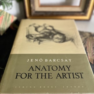 Könnte beinhalten: Ein Vintage-Buch mit dem Titel "Anatomie für den Künstler" von Jenő Barcsay. Das Buch hat einen hellgrünen und cremefarbenen Einband mit schwarzem Text und einer Skizze einer Figur. Das Buch ist auf einer gemusterten Oberfläche geöffnet.