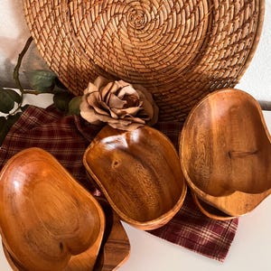 Pode incluir: Três tigelas de madeira com acabamento natural são dispostas sobre um tecido xadrez vermelho e branco. Um prato grande tecido e uma rosa decorativa estão no fundo. As tigelas têm uma aparência lisa e polida e formas variadas.