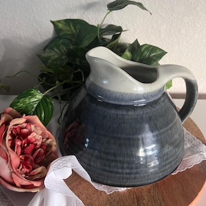 May include: A ceramic pitcher with a dark blue glaze and a light gray rim. The pitcher has a handle and a spout. It sits on a wooden tray with a white lace doily. A pink rose and green leaves are in the background.