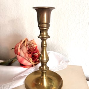 May include: A vintage brass candlestick holder with a wide base and a slender, ornate stem. The candlestick has a flared top and a warm, golden hue. A faded pink rose and white lace are in the background.