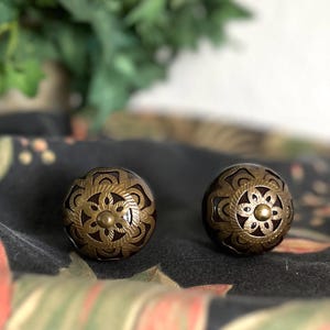 Puede incluir: Dos pomos redondos para armarios con una base de color marrón oscuro y un intrincado revestimiento metálico en relieve de color latón. El diseño metálico muestra un patrón floral simétrico con un perno circular central y formas de pétalos.