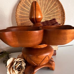 Pode incluir: Uma travessa de servir de madeira vintage com três tigelas e um elemento decorativo central. A travessa é feita de madeira polida com um tom castanho quente. Um prato de vime tecido e uma rosa seca estão no fundo.