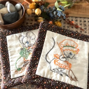 May include: Two square, embroidered fabric coasters. Each coaster features a white square with a whimsical mouse design. One mouse knits, the other sweeps. The coasters have a dark brown floral border.