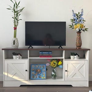May include: A white and brown TV stand with a black flat-screen TV on top. The stand has two cabinet doors with an X design, and open shelving. Decorative items include plants, books, and a vase of flowers.