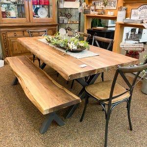 May include: A wooden dining table with a live edge design, paired with a matching bench and chairs. The table has a natural wood finish and black metal legs. The chairs have brown leather seats.