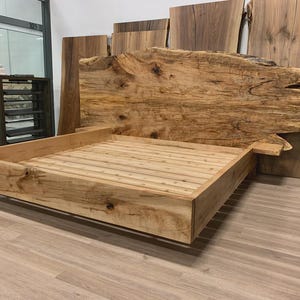 May include: A handcrafted wooden bed frame with a natural, unfinished appearance. The headboard is a large, live-edge slab, and the bed frame has a simple, modern design. The bed frame is made of light-coloured wood.