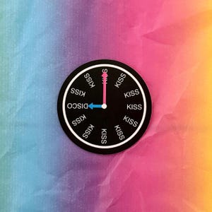 May include: A black circular sticker with a clock design. The clock face has the word "DISCO" and the word "KISS" repeated around the perimeter. The clock hands point to "S" and "DISCO". The background is a gradient of blue, pink, and yellow.