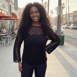 May include: A black crochet sweater with a mesh design on the upper chest and sleeves. The sweater has long sleeves and a round neckline. The person is wearing black pants. The background shows a street with palm trees and buildings.