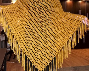 Peut inclure: Un poncho au crochet jaune moutarde présentant un motif de mailles ajourées en forme de losanges. Le vêtement possède un ourlet en pointe agrémenté de longues franges en fil pendantes sur tout le bord inférieur, créant un aspect texturé.