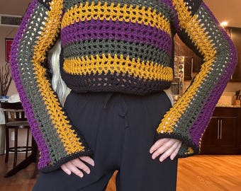 Op de afbeelding: Een handgemaakte gehaakte trui in een cropped stijl. De trui heeft horizontale strepen in zwart, goud, paars en olijfgroen. De mouwen zijn lang en klokvormig. De trui wordt gecombineerd met een zwarte broek.