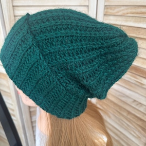 May include: A dark forest green beanie hat crafted with a textured crochet stitch pattern. The hat features a slouchy silhouette with a ribbed band at the base and a soft, slightly fuzzy yarn texture throughout the entire exterior surface.