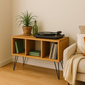 May include: A light oak record player stand with black hairpin legs. The stand has three cubby holes, one with green books and another with vinyl records. A black record player sits on top, along with a potted plant and a green bottle.
