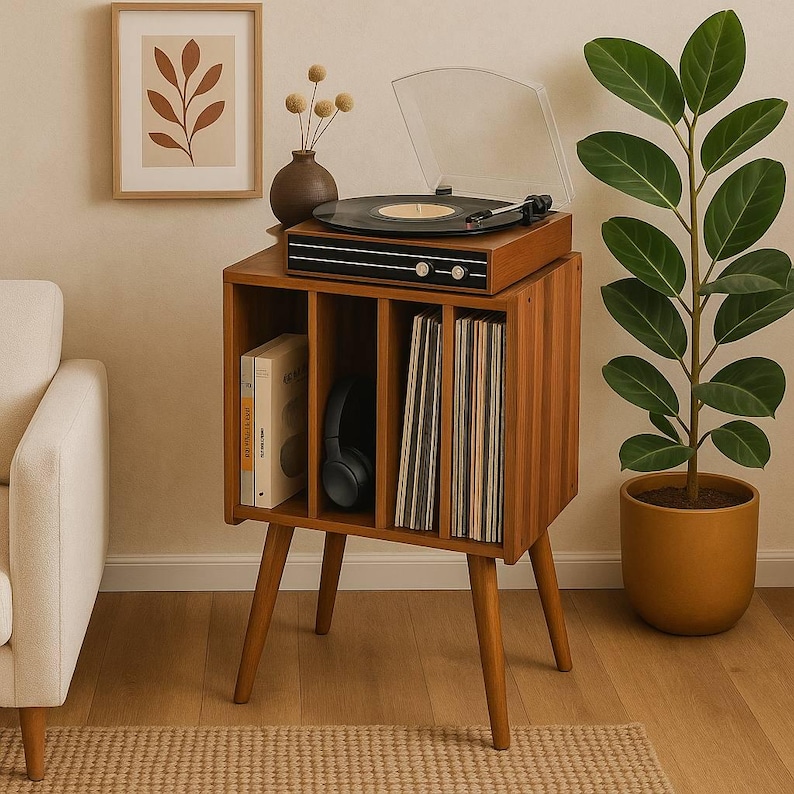 May include: A mid-century modern record player and storage cabinet in a warm wood tone. The turntable is playing a record, with a clear dust cover. The cabinet has compartments for vinyl records, books, and headphones. A framed botanical print and potted plant complete the scene.