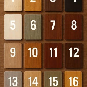 May include: Sixteen wooden square tiles with different wood stain colors, numbered 1 through 16 in white. The tiles are arranged in a grid pattern on a wooden surface, showcasing a variety of hues from light beige to dark brown.
