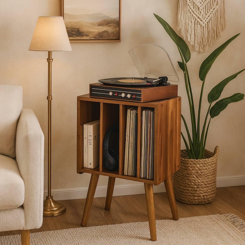 May include: A wooden record player stand with a built-in record storage compartment. The stand has a vintage record player on top, with a clear plastic cover. The stand is made of light brown wood and has four angled legs. A floor lamp and a houseplant are also in the image.