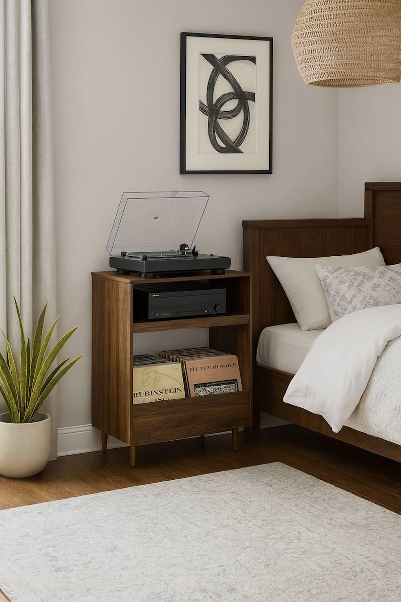 May include: A wooden record player stand with a turntable, amplifier, and record storage. The stand is next to a bed with white bedding and a framed abstract art piece. A potted plant and rug are also in the room.