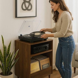 May include: A woman placing a vinyl record on a black turntable atop a dark wood record cabinet. The cabinet has a shelf with a stereo receiver and a lower shelf with vinyl records. A framed abstract art piece hangs on the wall.