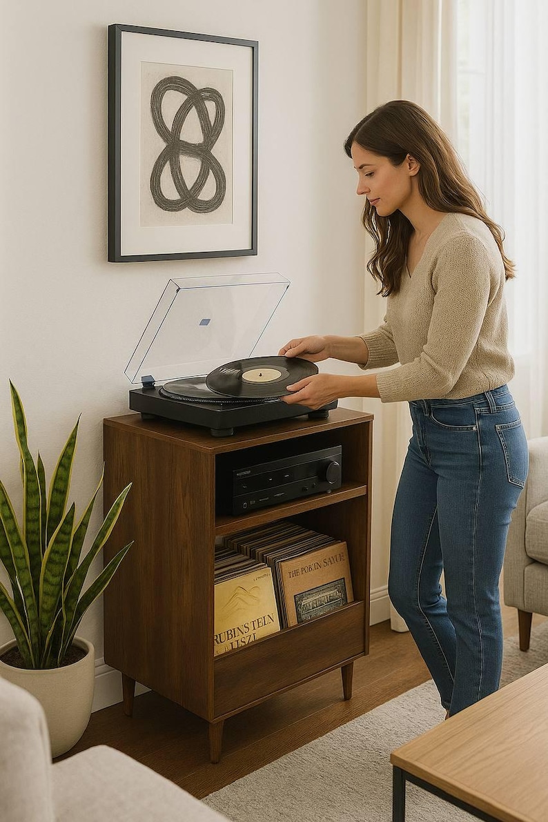 May include: A woman placing a vinyl record on a black turntable atop a dark wood record cabinet. The cabinet has a shelf with a stereo receiver and a lower shelf with vinyl records. A framed abstract art piece hangs on the wall.