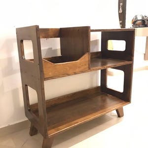 May include: A dark brown wooden bookshelf with two shelves and a small cubby. The sides feature rectangular cutouts. The bookshelf stands on four angled legs. The unit is designed for storage and display.