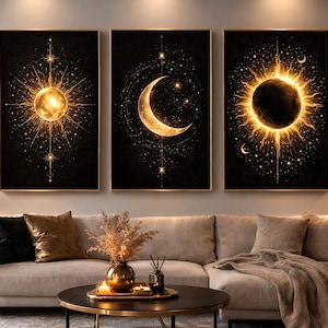 May include: Three framed celestial art prints. Each features a gold sun, moon, or sunburst design on a black background. The artwork is displayed above a beige sofa with decorative pillows and a blanket. A gold lamp and plants complete the room.