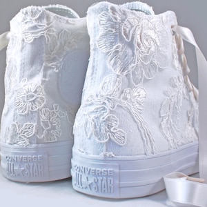 May include: White high-top sneakers with white lace and a white satin ribbon. The sneakers are decorated with white floral embroidery and the words "Converse All-Star" are printed on the side.
