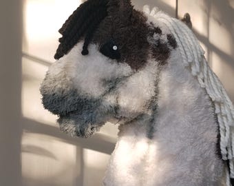 May include: A plush hobby horse toy featuring a white and brown textured fabric body. The horse has a dark brown forelock, a long white yarn mane, and a dark eye with a white center. The muzzle area is a mix of white and grey plush material.