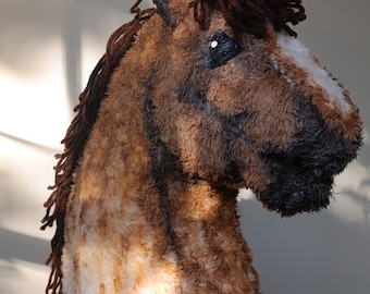 May include: A plush horse head toy featuring a textured brown and tan coat with a white blaze on the nose. The horse has a dark brown yarn mane, black yarn muzzle detailing, and black eyes with white accents, crafted from soft, fuzzy material.