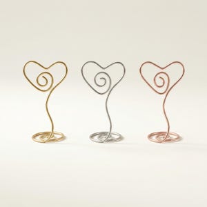 May include: Three heart-shaped wire holders in gold, silver, and rose gold. Each holder features a heart design with a spiral detail and a coiled base. The holders are displayed against a white background.