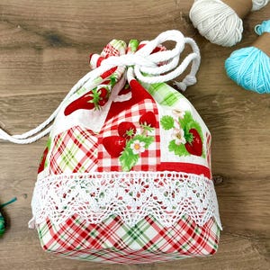 Puede incluir: Una bolsa con cordón que presenta un diseño de patchwork de fresas rojas, hojas verdes y flores blancas sobre un fondo de cuadros rojos y blancos, acentuada con un borde de encaje blanco y cordones gruesos blancos.