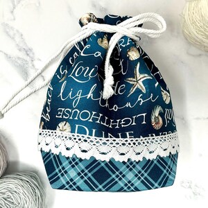 May include: A drawstring pouch featuring a dark teal fabric with white cursive text reading lighthouse, beach, and dune, accented by starfish and seashell illustrations. The bottom section is a teal plaid pattern separated by white lace trim.