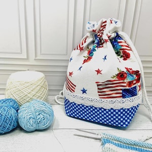 May include: A drawstring bag featuring a white fabric with a red, white, and blue American flag and floral pattern, accented by a white lace trim and a blue and white gingham base. The bag includes thick white rope drawstrings for closure.