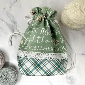 May include: A sage green drawstring pouch featuring a white nautical print with seashells and text including Tide and Lighthouse. The bag has a white lace trim separating the top section from a bottom panel with a white, green, and brown plaid pattern.