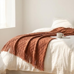 Pode incluir: Uma manta de malha cor ferrugem com um padrão de losangos e detalhes de franjas está sobre uma cama. Uma caneca branca está em um livro na cama. A cama tem lençóis brancos.