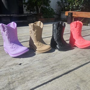 May include: Four miniature cowboy boots in a row, each a different color: lavender, tan, black, and coral. The boots have detailed embossed designs and are arranged on a weathered wooden surface.