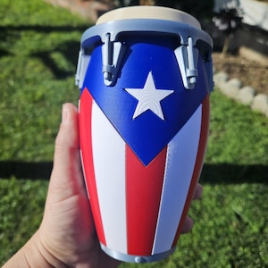May include: A small, decorative conga drum featuring the Puerto Rican flag design. The drum has a blue top with a white star, red and white stripes, and a light tan drumhead. The drum is held in a hand against a blurred green background.