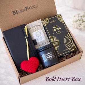 Puede incluir: Caja de regalo "BlissBox" con un cuaderno negro, bol&iacute;grafo dorado, sobre de t&eacute;, tableta de chocolate negro, vela de vainilla y un coraz&oacute;n rojo tejido. La caja tiene el texto "Bold Heart Box".