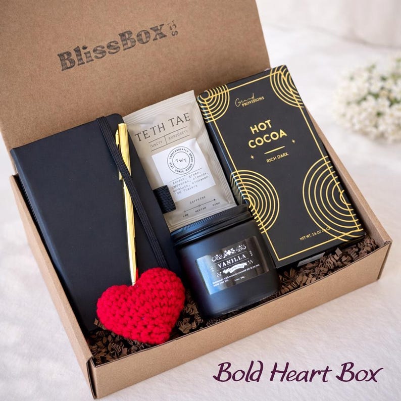 Puede incluir: Caja de regalo "BlissBox" con un cuaderno negro, bol&iacute;grafo dorado, sobre de t&eacute;, tableta de chocolate negro, vela de vainilla y un coraz&oacute;n rojo tejido. La caja tiene el texto "Bold Heart Box".