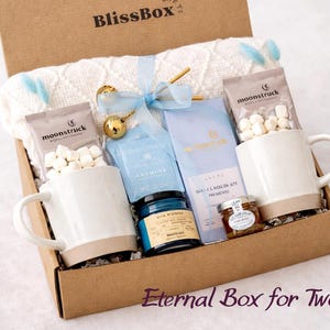 May include: A cardboard gift box labeled BlissBox containing two speckled ceramic mugs, two packets of Moonstruck marshmallows, a white knit blanket with blue accents, a blue tea tin, a blue box, a small jar of honey, and a gold tea infuser.