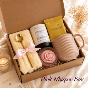 May include: A gift box with a pink theme, containing a mug, tea, a candle, a spoon, and a soft blanket. The box includes a candle shaped like a rose and a gold spoon. The text "Pink Whisper Box" is visible.