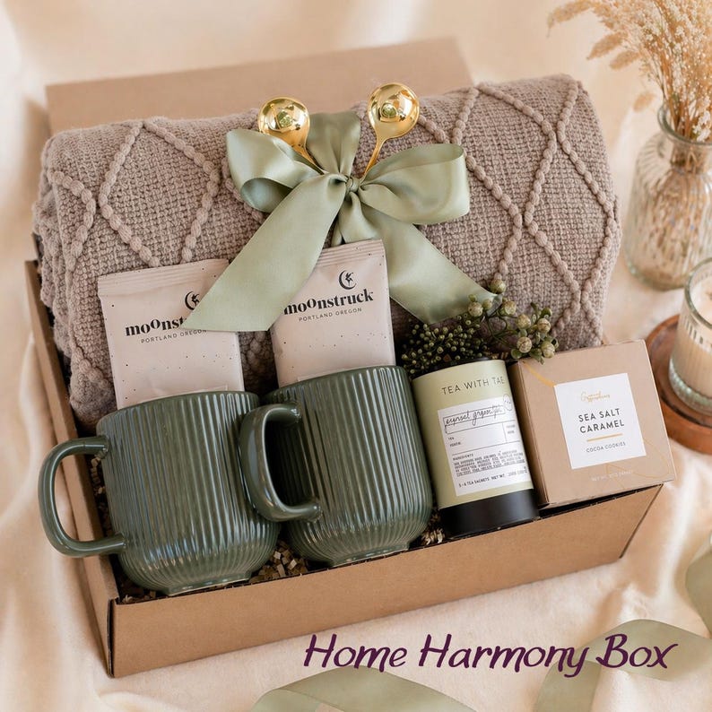 May include: A gift box containing a taupe textured throw blanket tied with a sage green ribbon and two gold spoons, two olive green ribbed ceramic mugs, two Moonstruck chocolate bars, a Tea with Tae tin, and a box of Sea Salt Caramel cookies.