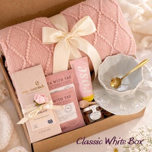 Op de afbeelding: Een geschenkdoos met een gevouwen oudroze gebreide deken vastgebonden met een cr&egrave;mekleurig lint, een Moonstruck chocoladereep, twee Tea With Tae zakjes, een tube met rozenlabel, een honingpotje met lepel en een witte theekop.