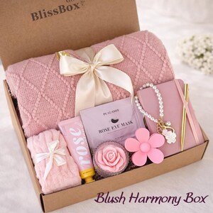 May include: A gift box labeled "BlissBox" filled with pink items. Includes a blanket, a notebook, a bracelet, a flower-shaped item, a rose-shaped soap, a tube of rose-scented lotion, and a "Rose Eye Mask" packet. The box is labeled "Blush Harmony Box".