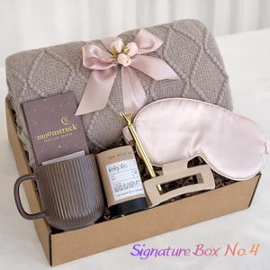 Könnte beinhalten: Eine Geschenkbox mit einer taupefarbenen, strukturierten Strickdecke mit rosa Satinschleife, einer Moonstruck Portland Oregon Schokolade, einem gerippten braunen Becher, einer Tea With Tae Teedose, einem goldenen Stift, einer Haarspange und einer hellrosa Schlafmaske aus Satin.