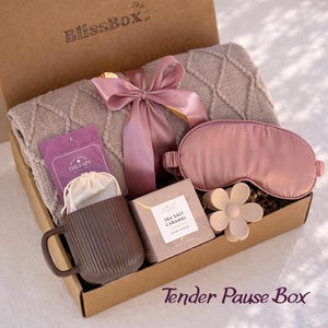 May include: A gift box containing a beige cable-knit blanket tied with a mauve satin ribbon, a mauve sleep mask, a brown ribbed ceramic mug, a small white drawstring pouch, a flower-shaped hair clip, and a box labeled Sea Salt Caramel.